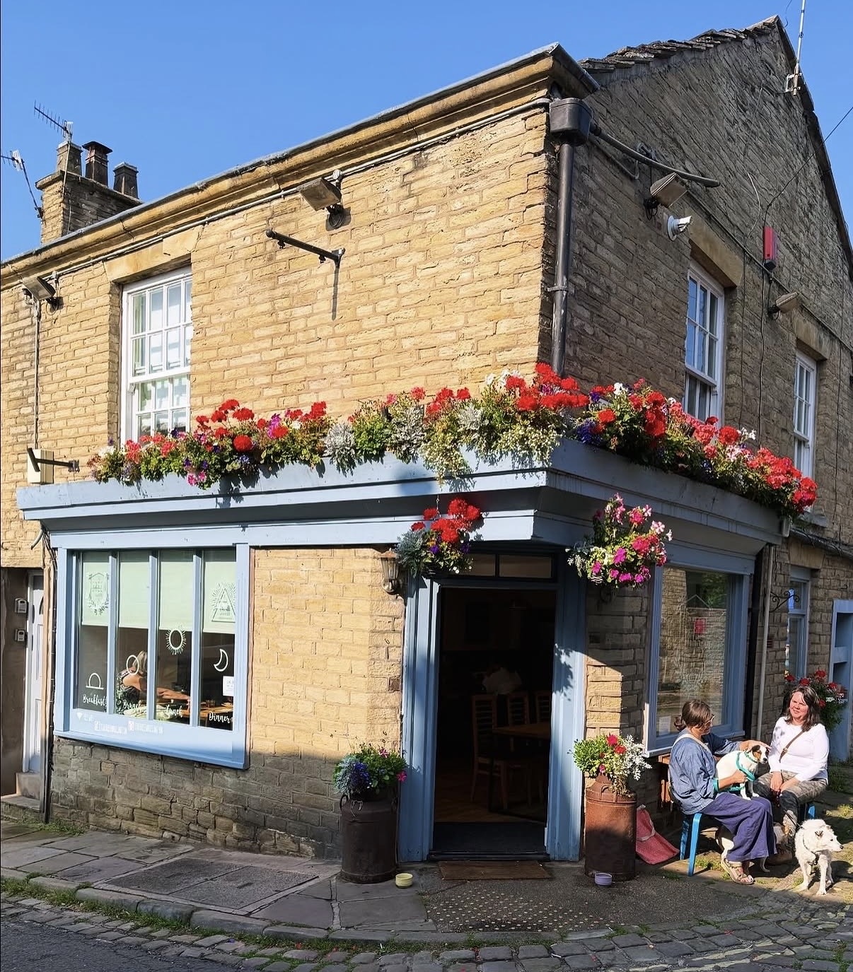 The Green's stone corner building in sunshine, with window boxes of red geraniums and customers with dogs sat outside